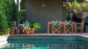 Una zona de piscina con borde de azulejos, sillas de madera y plantas en macetas.