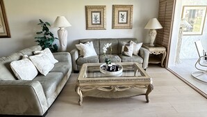 A living room with a beige sofa, a glass coffee table, and a ceiling fan. There are two framed pictures on the wall and a plant on the side. - GOLF INCLUDED!! Charming 1st Floor Condo; 1.5 miles to Naples’ Beaches (Naples)