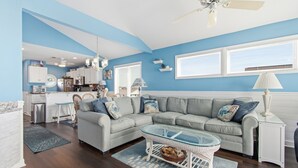 A living room with a grey sectional sofa, a glass coffee table, and a white side table. There is a kitchen in the background with white cabinets and a window. - "Waters Edge 427-A" - Oceanfront Condo w/ Community Pool, 2 Bedrooms / 2 Bathrooms (Holden Beach)
