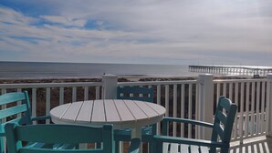 A deck with a round table and chairs, overlooking a beach and a pier. - "Waters Edge 427-A" - Oceanfront Condo w/ Community Pool, 2 Bedrooms / 2 Bathrooms (Holden Beach)