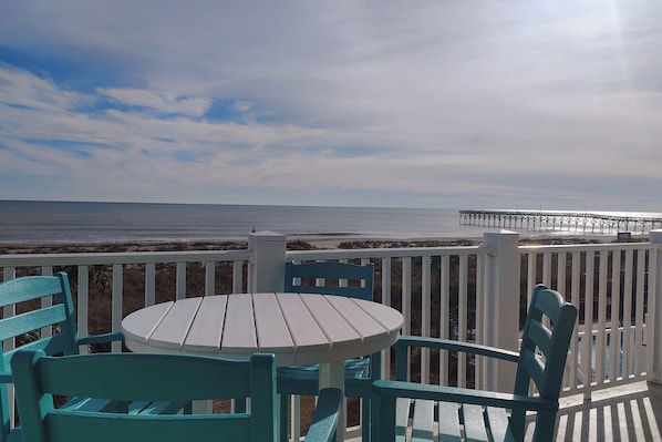 A deck with a round table and chairs, overlooking a beach and a pier. - "Waters Edge 427-A" - Oceanfront Condo w/ Community Pool, 2 Bedrooms / 2 Bathrooms (Holden Beach)