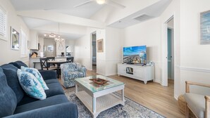 A living room with a sofa, coffee table, and TV. There is a dining area with a table and chairs. The room has white walls and a ceiling fan. - "Waters Edge 427-B" - Oceanfront Condo w/ Community Pool, 2 Bedrooms / 2 Bathrooms (Holden Beach)