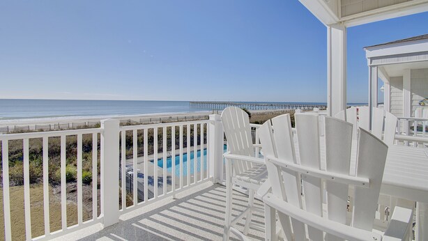A balcony with white chairs and a pool, overlooking a beach and pier. - "Waters Edge 427-B" - Oceanfront Condo w/ Community Pool, 2 Bedrooms / 2 Bathrooms (Holden Beach)