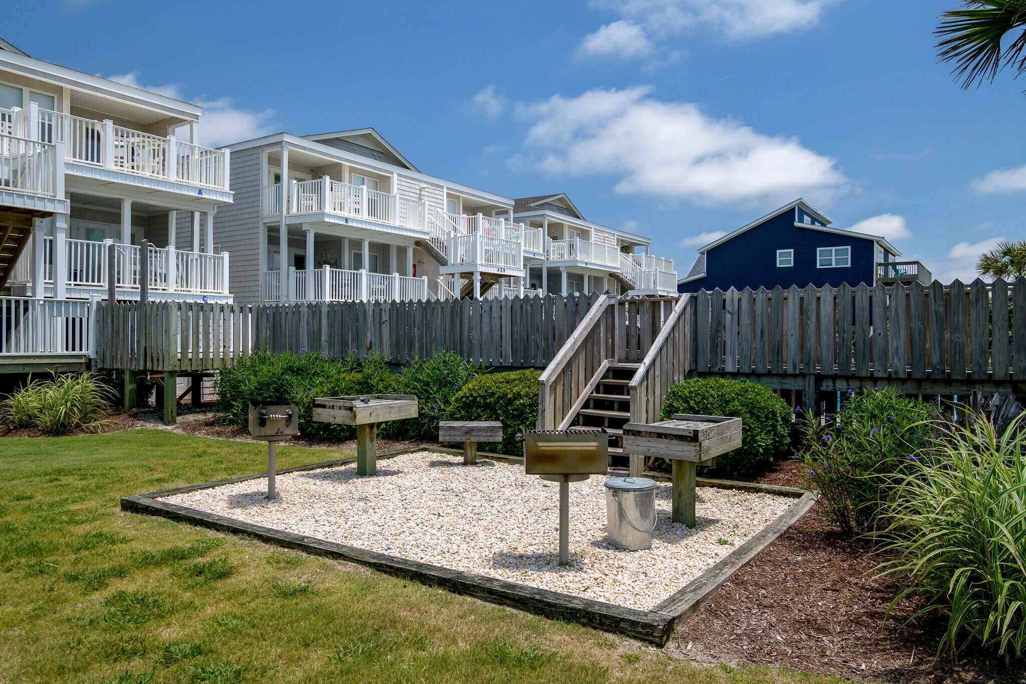 A beachfront property with multiple-story buildings, a wooden fence, and a landscaped garden with benches and mailboxes.