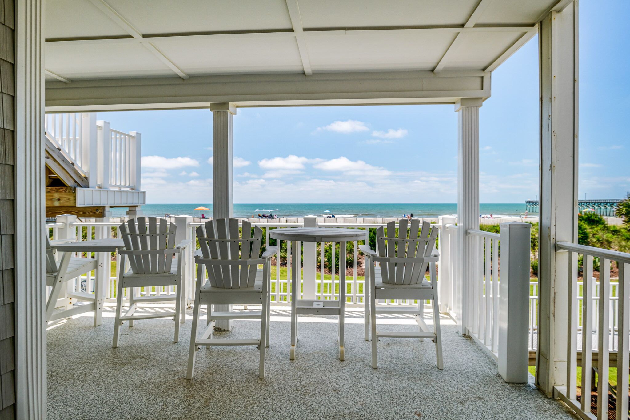 A covered outdoor seating area with a table and four chairs, overlooking a beach with a pier in the distance.