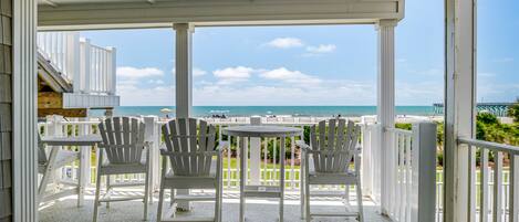 A covered outdoor seating area with a table and four chairs, overlooking a beach with a pier in the distance.