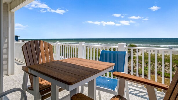 Une table et des chaises en bois sur un balcon doté d’une balustrade blanche, avec vue sur une plage où l’on aperçoit des vagues et un ciel dégagé.