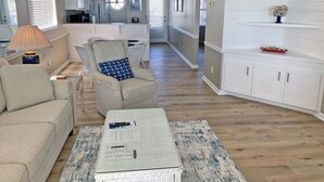 Un salon spacieux avec un canapé, un fauteuil et une table basse. On aperçoit une cuisine à l’arrière-plan, avec des meubles blancs et un luminaire suspendu.