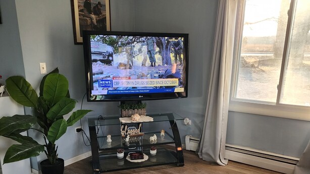 Ein Wohnzimmer mit Fernseher auf einem Schrank, einer Topfpflanze, einem Glastisch und einem Fenster mit Vorhängen.