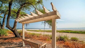 Eine Holzschaukel mit Metallgestell, eingebettet in eine natürliche Umgebung mit Bäumen und einem Strand im Hintergrund.