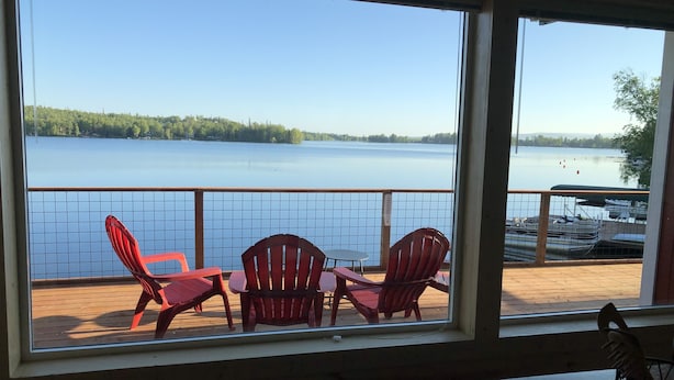 A room with a large window overlooking a lake, featuring a wooden deck with red chairs and a table. - Cozy Lakefront Cabin on Big Lake, Alaska (Big Lake)