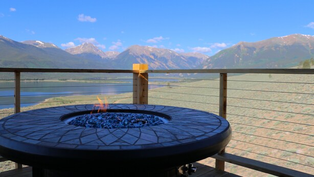 Una griglia circolare da esterno con un fuoco, posizionata su un pontile in legno con vista su un lago e montagne.