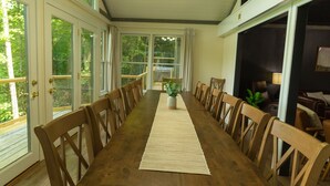 Ein langer Holztisch mit Stühlen, eine hängende Chandelierleuchtre und große Fenster mit Vorhängen.