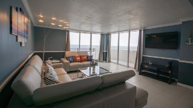 Un salon spacieux avec un grand canapé, une table basse et un téléviseur à écran plat fixé au mur. La pièce est dotée de grandes fenêtres qui offrent une vue sur la mer.