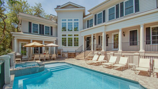 A swimming pool with lounge chairs and umbrellas, surrounded by a two-story building with large windows and a covered patio area. - Castle by the Sea 7BR/8BA Luxury Home with Elevator (Hilton Head Island)