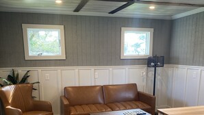 A room with a brown leather sofa, a wooden coffee table, and a white cabinet. There are two windows with white frames, and the walls have a paneling design. - Our Lucky Charm is a cottage on the waterway in Hatteras Village  (Hatteras)