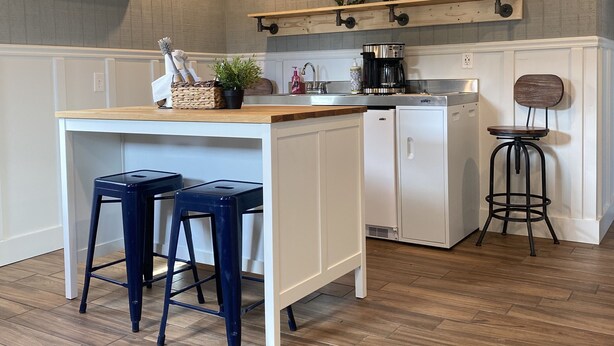 A kitchen with a central island, two blue bar stools, a coffee maker, and a hanging shelf with decorative items. - Our Lucky Charm is a cottage on the waterway in Hatteras Village  (Hatteras)