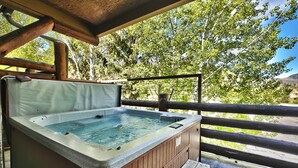 Ein Whirlpool mit Holzverkleidung und einer Sitzbank, umgeben von einer Holzdielen-Terrasse und einem Metallgeländer, mit Bäumen im Hintergrund.