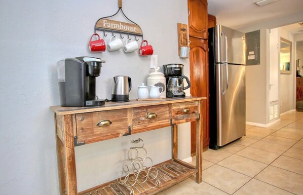 A kitchen with a wooden console table holding coffee makers, a kettle, and mugs. A stainless steel refrigerator is to the right.