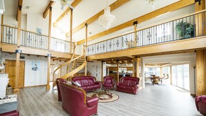 En rummelig opholdsstue med højt til loftet, træbjælker og en trappe, der fører til en etage ovenpå. Der er flere lædersofaer og en lysekrone.