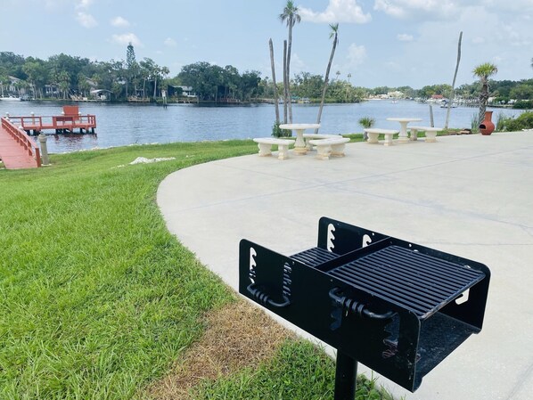 A waterfront park with a barbecue grill, picnic tables, and a red dock.