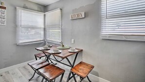 Un comedor con una mesa de madera y bancos, dos ventanas con persianas, y un cartel en la pared que dice 'KITCHEN'.