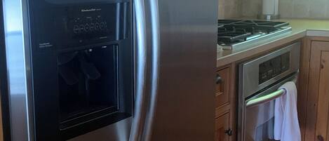 A stainless steel refrigerator with a digital display, a microwave, and a stove in a kitchen with wooden cabinets and a window.