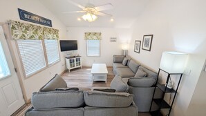 A living room with a sofa, a TV, a ceiling fan, and a sign that reads 'BEACH HOUSE'. - West Side Near Avalon Pier ~ Hot Tub~Dog Friendly~Firepit + Fenced in Yard (Kill Devil Hills)