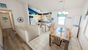 A kitchen with wooden cabinets, a central island, and a dining area with a table and chairs. - West Side Near Avalon Pier ~ Hot Tub~Dog Friendly~Firepit + Fenced in Yard (Kill Devil Hills)