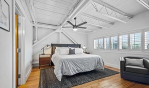 A bedroom with a bed, a nightstand, a desk, a chair, a ceiling fan, and large windows.