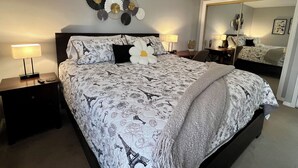 Une chambre à coucher bien aménagée, avec un grand lit, deux tables de chevet avec des lampes, un miroir et un élément décoratif mural au-dessus du lit.