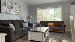 Un salon avec un canapé gris, un divan à motifs, une table basse blanche et une fenêtre avec des stores.