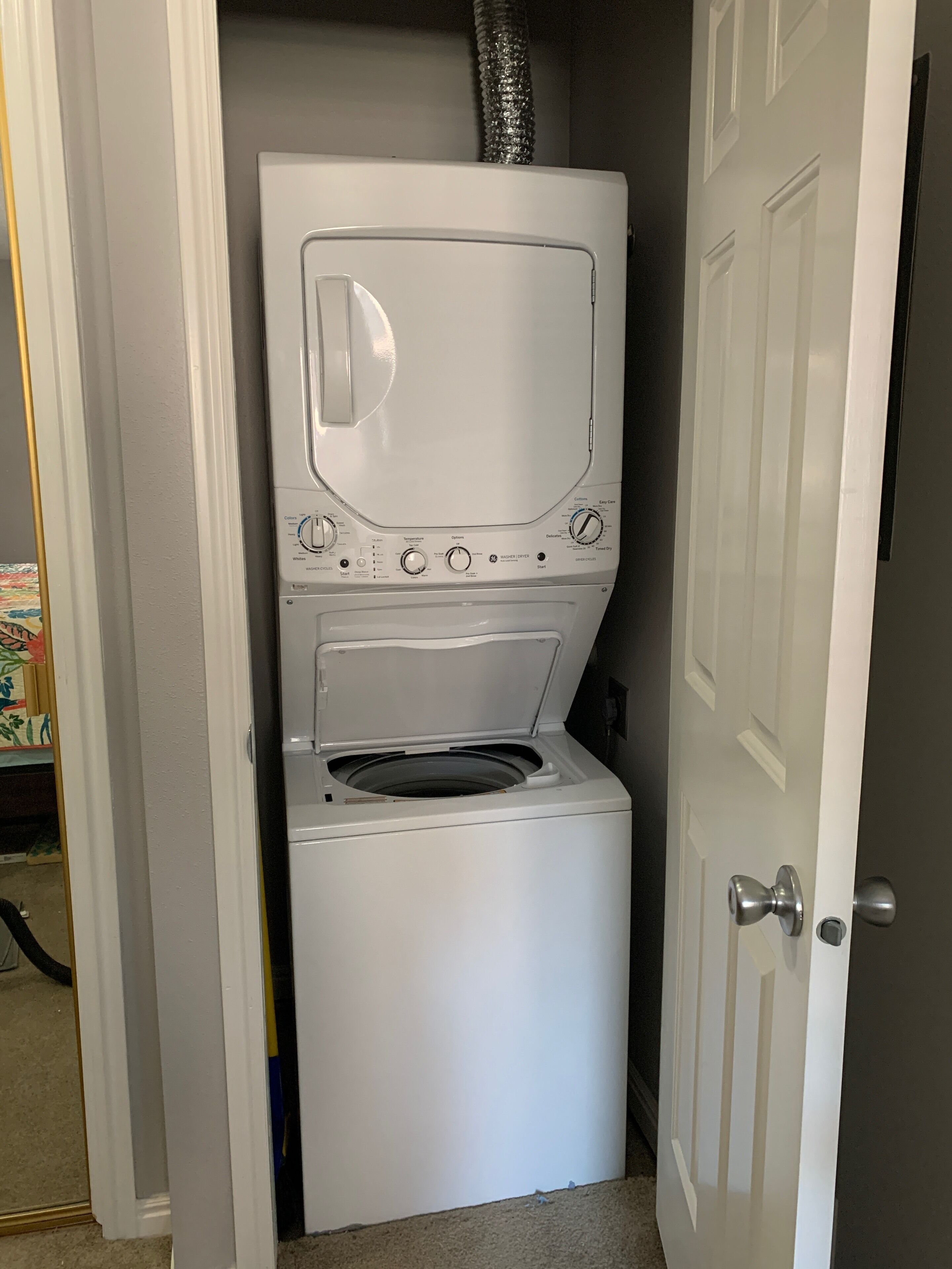 Un appareil compact, blanc, combinant une machine à laver et un sèche-linge à chargement frontal, installé dans un petit espace buanderie avec une porte sur le côté droit.