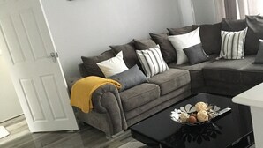 A living room with a grey sectional sofa, a black coffee table with decorative items, a grey rug, and a cityscape painting on the wall.