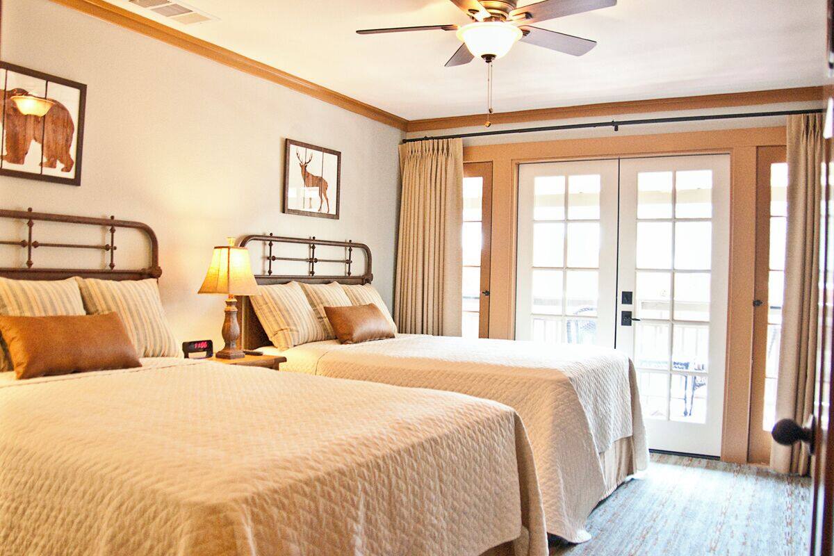 A hotel room with two beds, a ceiling fan, a nightstand with a lamp, and a large window with curtains.