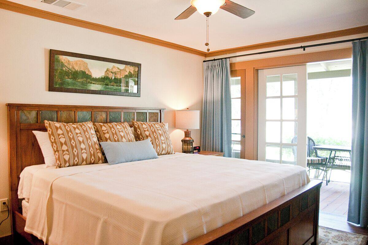 A large bed with patterned pillows, a ceiling fan, a painting of a landscape, and a view of a patio with outdoor furniture.