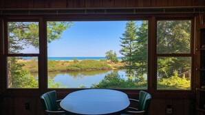 Una habitación con un amplio ventanal que da a un lago y un bosque. Hay una mesa redonda con dos sillas.