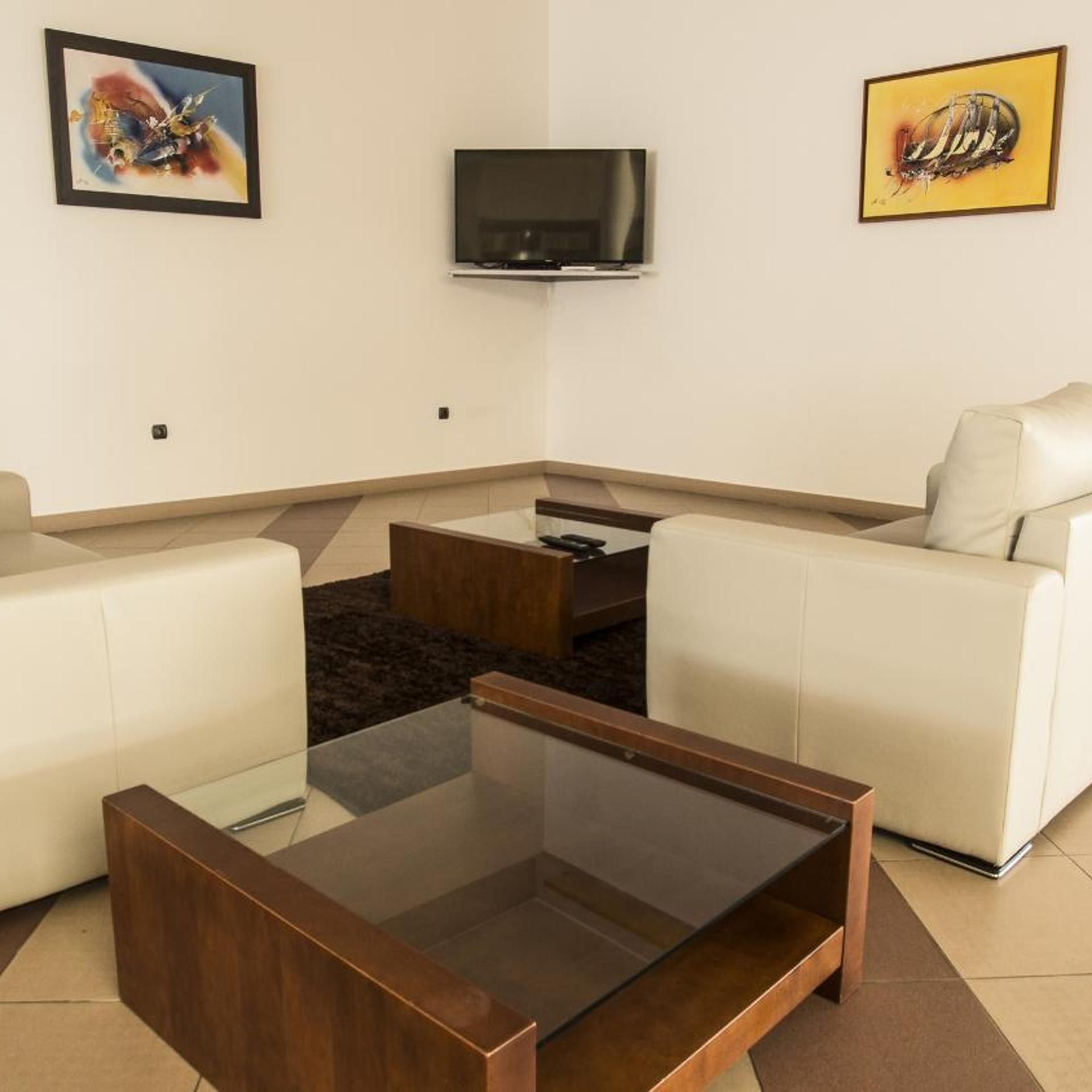 Un salon moderne avec une table basse en verre, deux canapés blancs, un téléviseur à écran plat et des œuvres d’art accrochées aux murs.