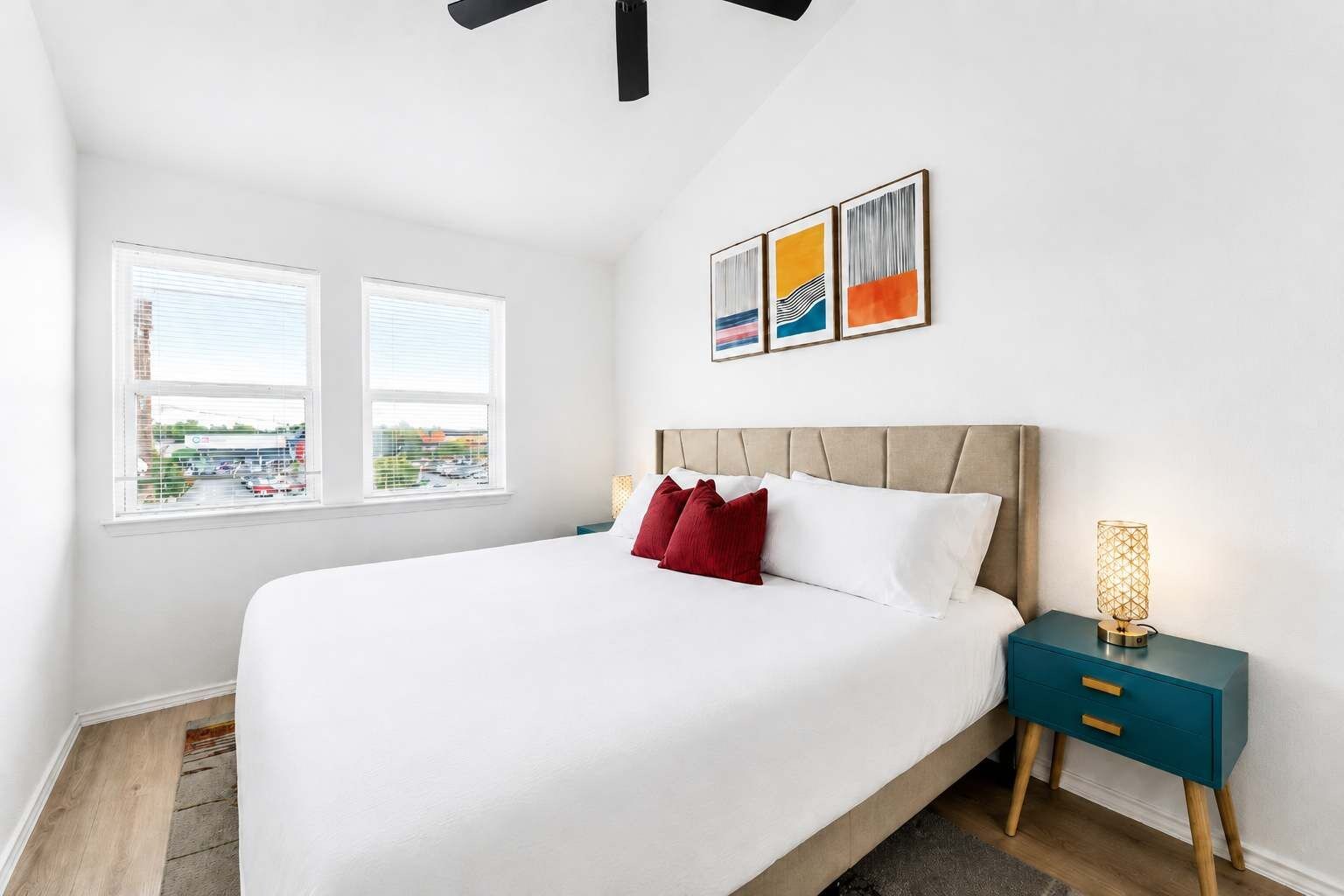 A bedroom with a bed, two windows, a nightstand, a lamp, and artwork on the wall.