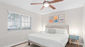 Una camera da letto con un letto, una ventola a soffitto, un comodino e una finestra con persiane.