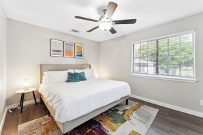 Relax in this serene bedroom with a large bed, wall-mounted fan, and window with horizontal blinds.