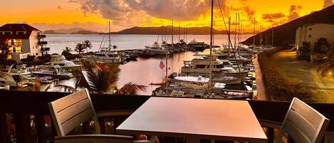 A view of a marina with boats docked, a table and chairs in the foreground, and a sunset in the background.