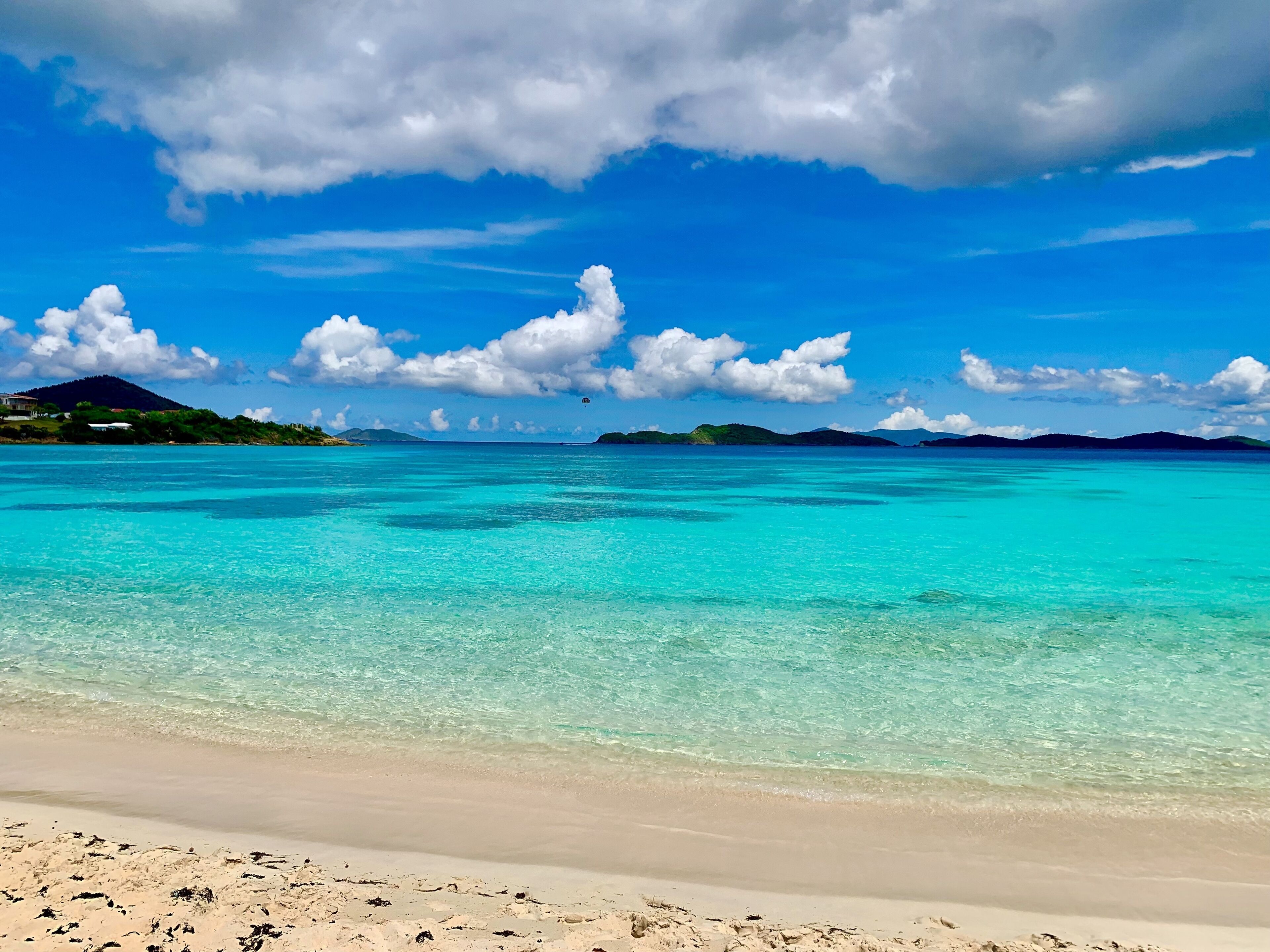 Une plage aux eaux turquoise et limpides, un rivage sablonneux et, au loin, des îles sous un ciel partiellement nuageux.