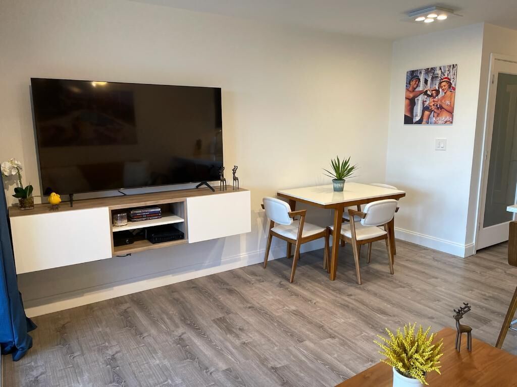 A modern living room with a flat-screen TV mounted on a white wall, a wooden dining table with chairs, and a potted plant on the table.