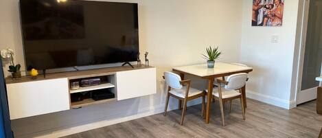 A modern living room with a flat-screen TV mounted on a white wall, a wooden dining table with chairs, and a potted plant on the table.