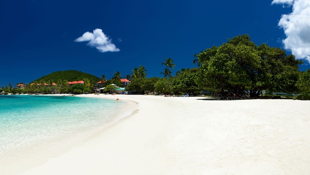 Une plage à l’eau turquoise et limpide, au sable blanc, avec une petite colline en arrière-plan, sur laquelle se dressent quelques bâtiments.