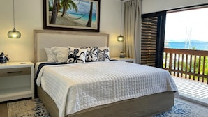 Une chambre avec un grand lit, un ventilateur de plafond, un tableau représentant une plage et un balcon avec vue.