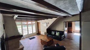 Ein geräumiges Wohnzimmer mit einer Holzbank, einem schwarzen Ledersofa und einem Fliesenboden. Eine Tür führt zu einem Balkon mit Blick auf Grünflächen.