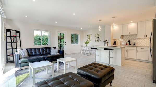 A modern living room with a black leather sofa, a white coffee table, and a black leather ottoman. There is a kitchen area with white cabinets and a herringbone tile backsplash.