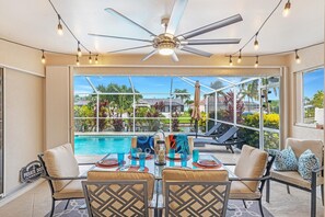 A poolside dining area with a table set for four, a ceiling fan, and a view of a swimming pool and tropical garden. - Direct Gulf Access Canal Home | Heated Pool, Game Room Murphy Bed & Fireplace | Sleeps 8 (Cape Coral)